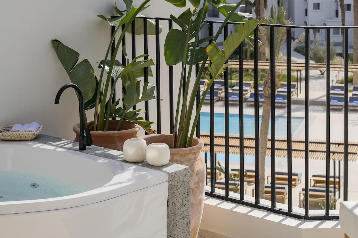 A balcony with a hot tub, potted plants, and a view of a pool and buildings.