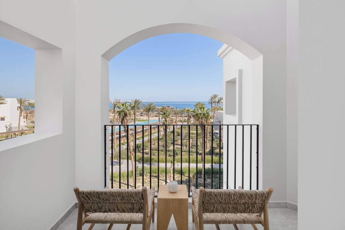 A balcony with two wicker chairs and a small table, overlooking a pool and palm trees.