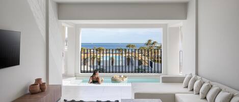 A modern living room with a large window overlooking a pool and ocean, a ceiling fan, and a flat-screen TV mounted on the wall.
