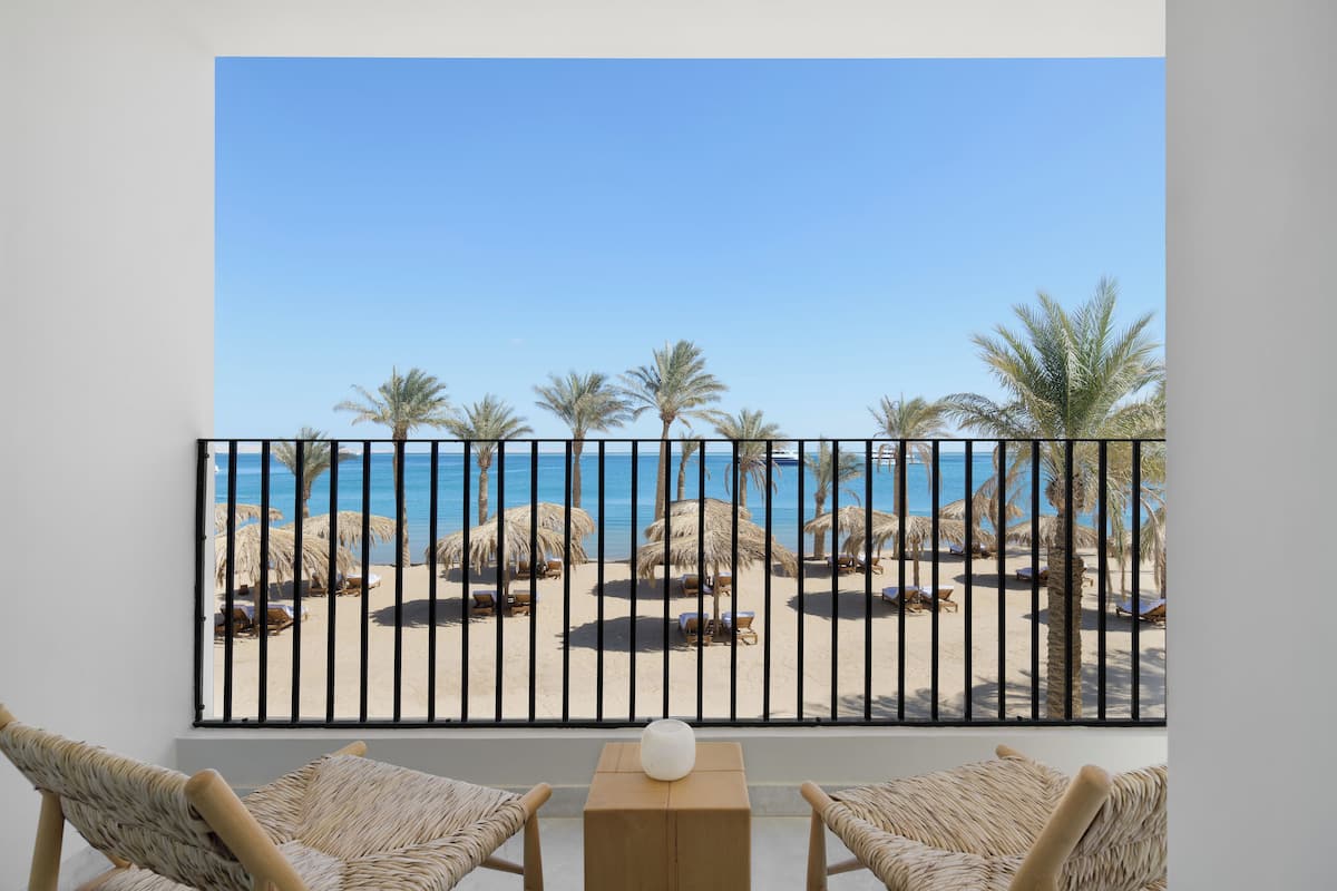 A balcony with wicker furniture, a wooden table, and a view of a beach with palm trees and lounge chairs.