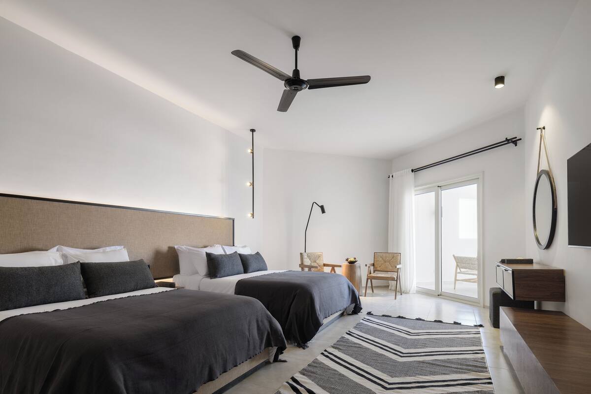 A modern hotel room with two beds, a ceiling fan, a large window, and a view of the outdoors.