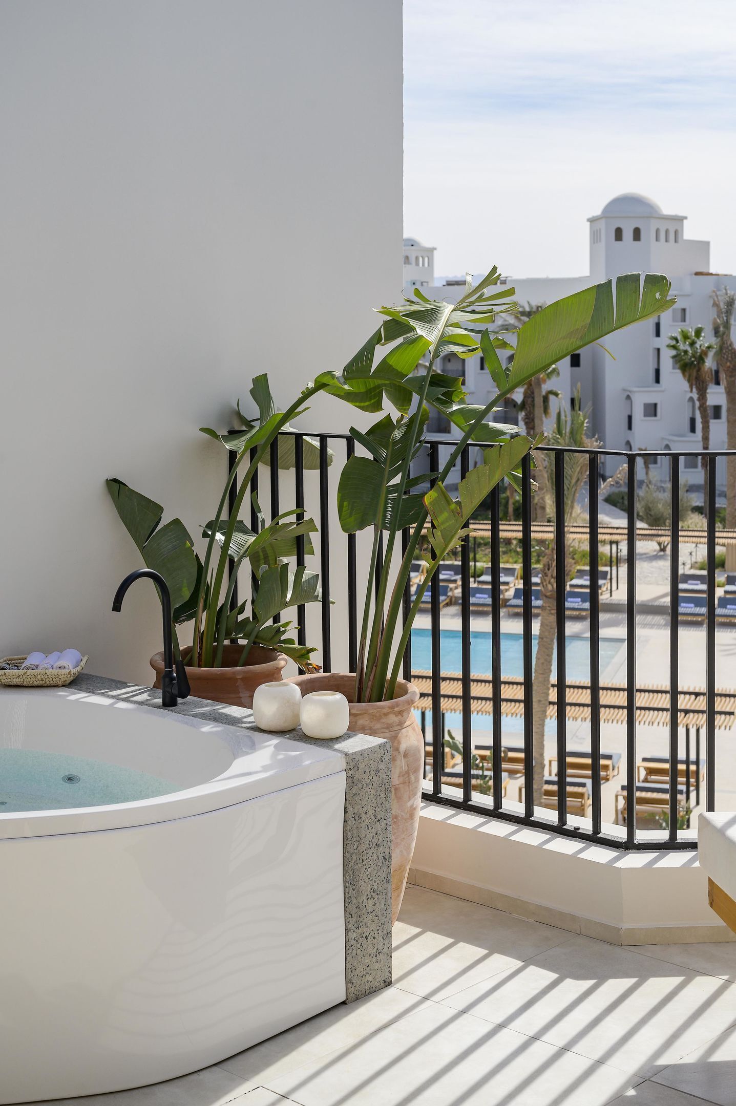 Un balcón con bañera de hidromasaje, plantas en macetas y vista a una piscina y edificios.