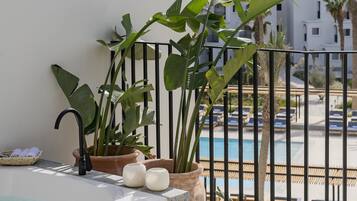 Un balcón con bañera de hidromasaje, plantas en macetas y vista a una piscina y edificios.