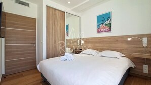 Un dormitorio con cabecera de madera, cama con ropa blanca, espejo de pared y un cartel de Cannes.