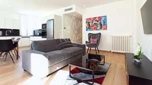 Una sala moderna con un sofá gris, una zona de comedor con una mesa y sillas, una cocina al fondo y un cuadro abstracto colorido en la pared.