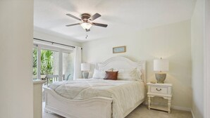 A bedroom with a bed, bedside tables, a ceiling fan, and a window with a view of greenery. - Beachy & Beautiful Renovated Villa Just Steps from (Hilton Head Island)