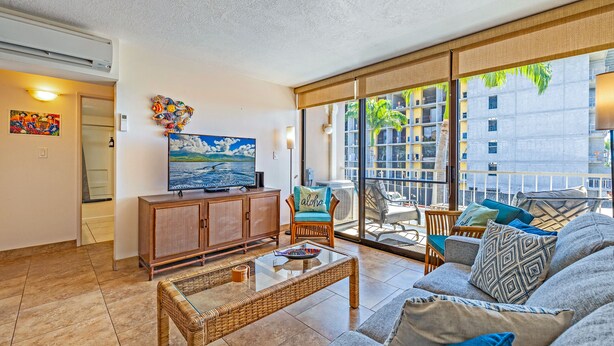 A spacious living room with a large television, wicker furniture, and a balcony with a view of buildings and palm trees. - Ocean View Sleeps 8 Across from Beach 3 bedroom 3 bathroom Full AC Kihei Condo  (Kihei)