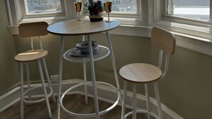 Una habitación con una mesa redonda, dos sillas, una planta en maceta y vistas a edificios a través de amplias ventanas.