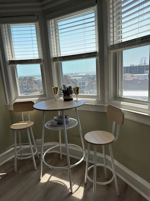 A room with a round table, two chairs, a potted plant, and a view of buildings through large windows.