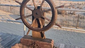 Una rueda de timón de madera sobre una plataforma de madera, con una playa arenosa y el océano al fondo.