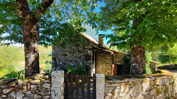 A rustic stone cottage with a wooden gate, surrounded by lush greenery and trees.