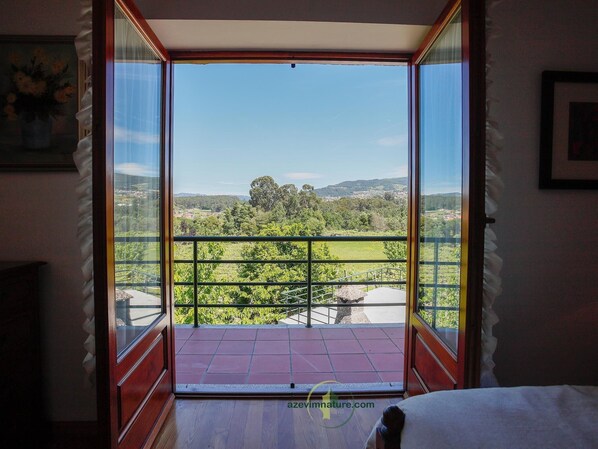 A room with a large glass door leading to a balcony with a metal railing, offering a view of a green landscape and distant hills.