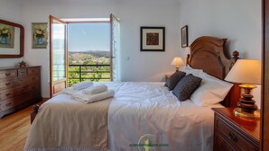 Una cama bien hecha con sábanas blancas y dos almohadas, un aparador de madera con cajones, una mesita de noche con lámpara y un cuadro en la pared.