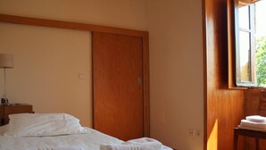 Een hotelkamer met een bed, een houten garderobe, een bureau met een lamp en een raam met uitzicht op groen.