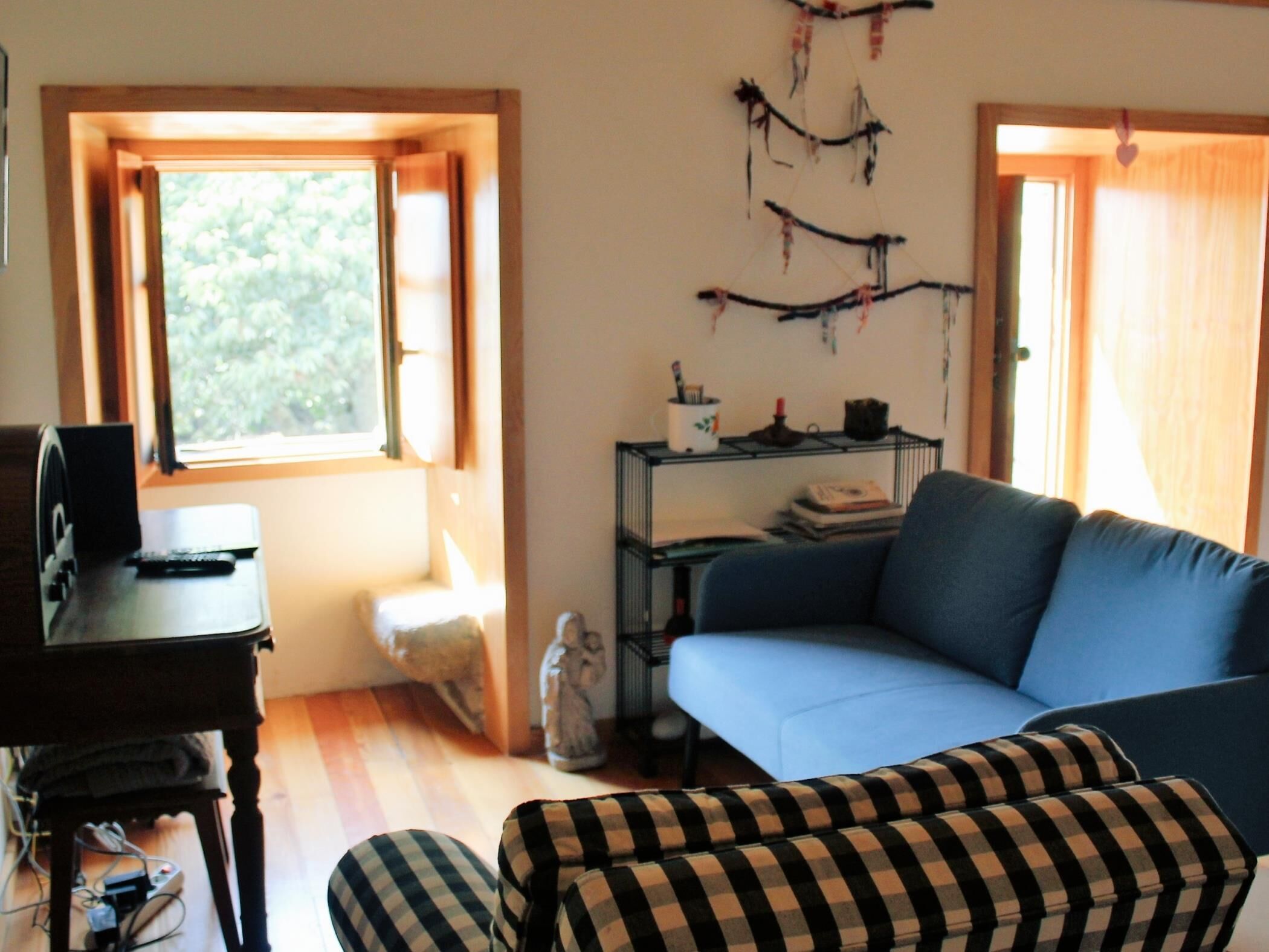 A living room with a blue sofa, a checkered ottoman, a small table with a remote control, and a wall-mounted shelf with books and decorative items.
