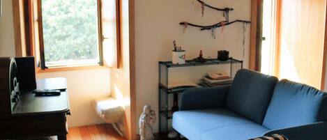 A living room with a blue sofa, a checkered ottoman, a small table with a remote control, and a wall-mounted shelf with books and decorative items.
