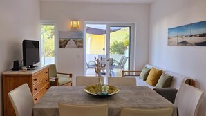 Ein Essbereich mit einem Tisch für vier Personen, einem Fernseher auf einer Holzkommode und Blick ins Freie durch eine Glastür.