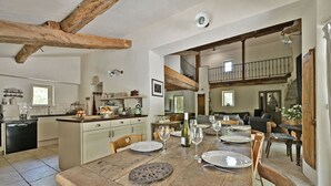 Een ruime eethoek met een houten tafel gedekt voor een maaltijd, een keuken met witte kasten en een slaapkamer op een entresol met een balkon.