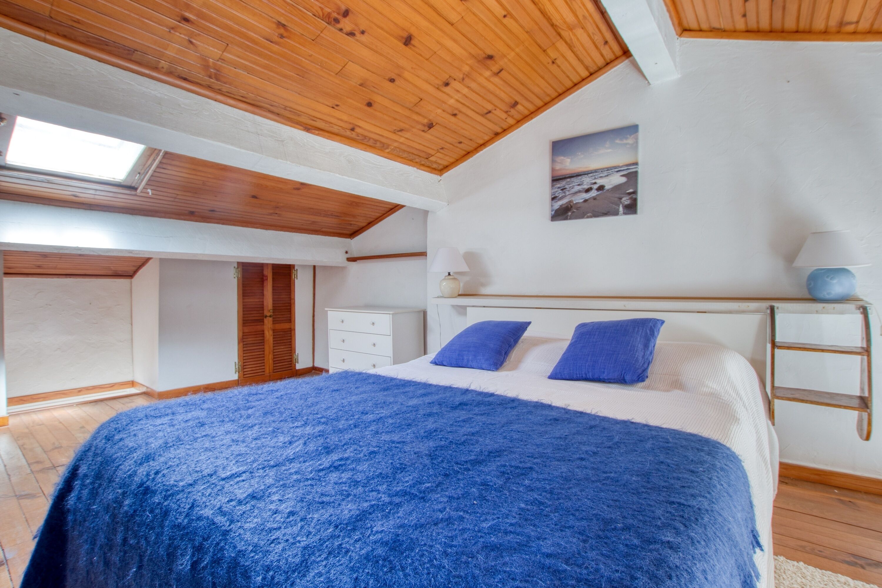 A bedroom with a large bed, a skylight, a white dresser, and a wooden ladder.