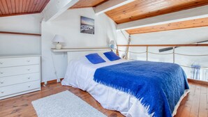 A bedroom with a wooden ceiling, a bed with a blue blanket, a white dresser, and a small bedside table with a lamp.