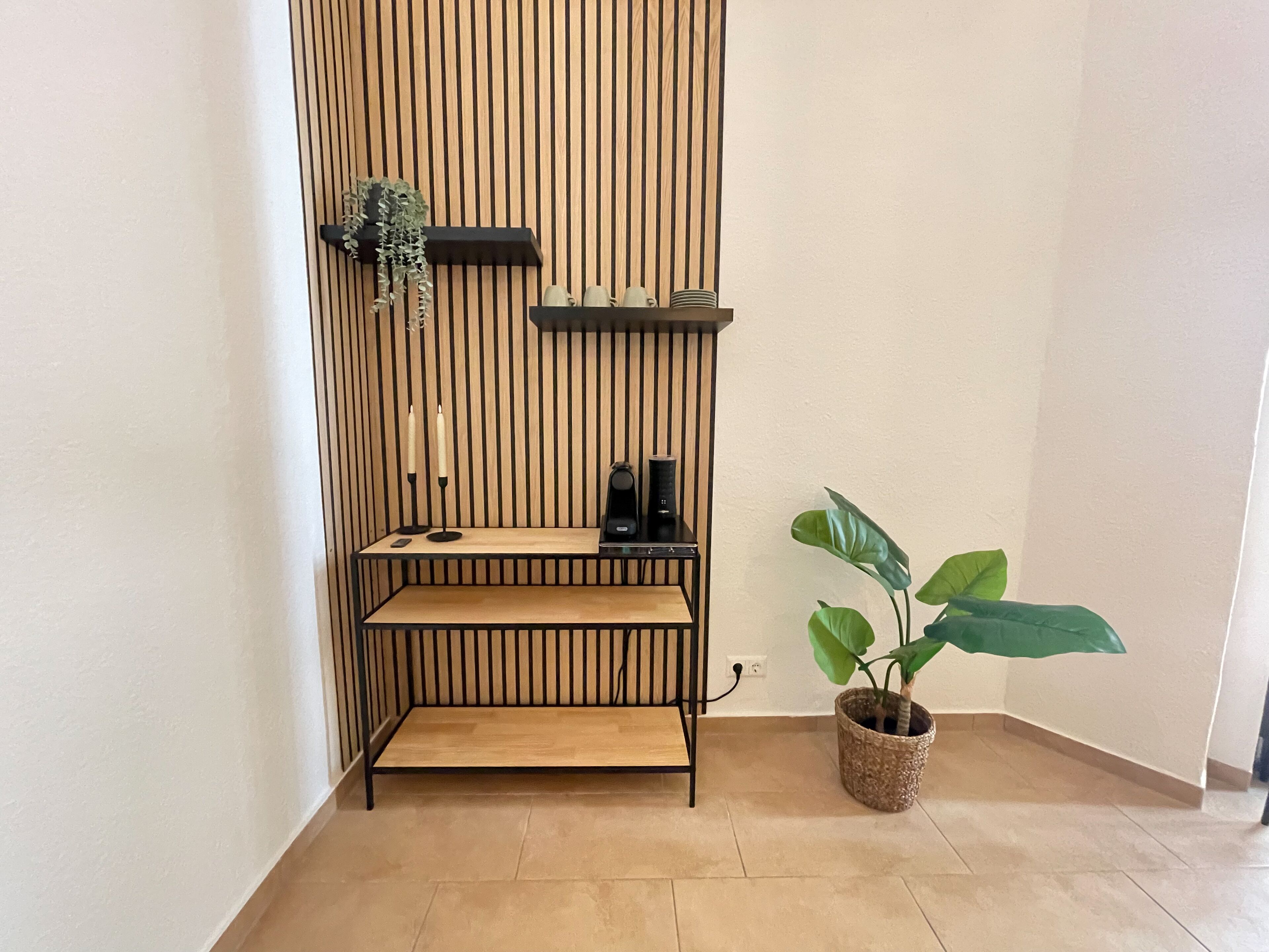 A corner with a wooden slat wall, a small bench, a shelf with candles and a diffuser, and a potted plant.