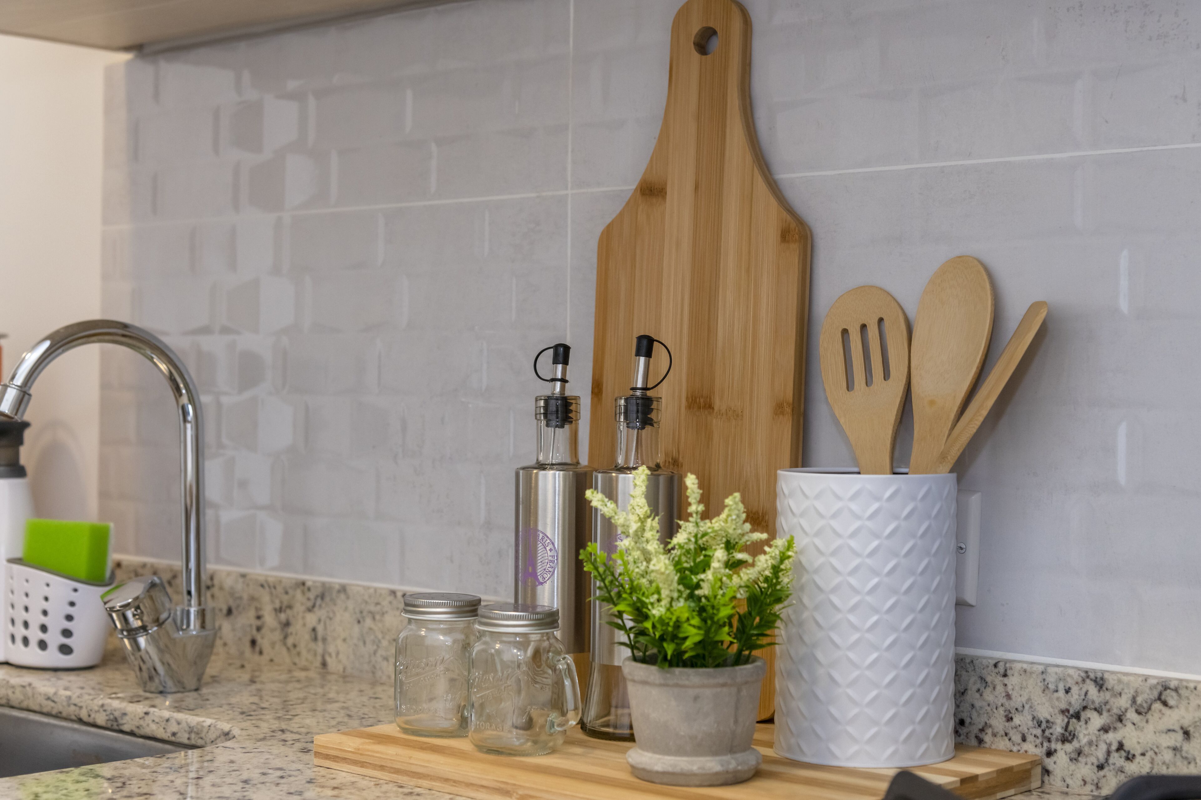 A kitchen counter with a sink, a wooden cutting board, a white container with wooden utensils, and a potted plant.