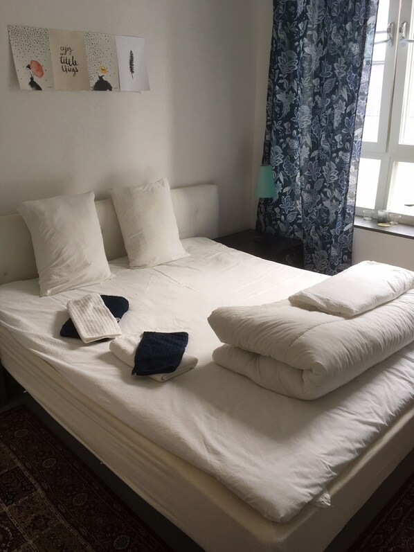 A neatly made bed with white linens and pillows, a folded towel, and a blue towel on the bed. There is a window with blue floral curtains and a lamp on the nightstand beside the bed.