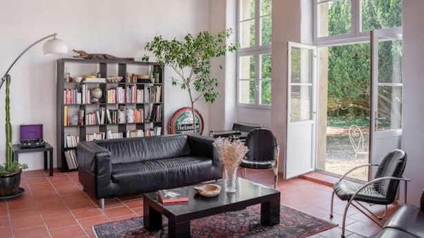 A spacious living room with a leather sofa, a coffee table, and a bookshelf. There are large windows providing a view of greenery outside. - Maison Familiale en Centre Historique (Talmont-Saint-Hilaire)