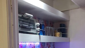 A compact kitchenette with a built-in stove, a sink, and a microwave. There are shelves above the counter holding various kitchen items.