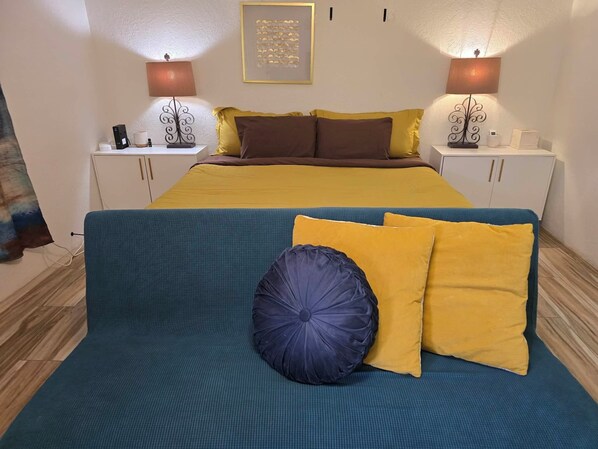 A bedroom with a bed, two bedside tables, a nightstand, a blue sofa, and a wall-mounted lamp. - Mirage- Private Secret Studio (Carlsbad)