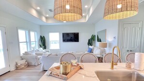 A spacious kitchen with a marble countertop, a sink, and a built-in dishwasher. There are wicker pendant lights, a dining area with a white table and chairs, and a large window providing natural light. - Crystal Cove Cottage - BEACHFRONT Luxury Getaway - Fully Fenced & Pet Friendly (Bolivar Peninsula)