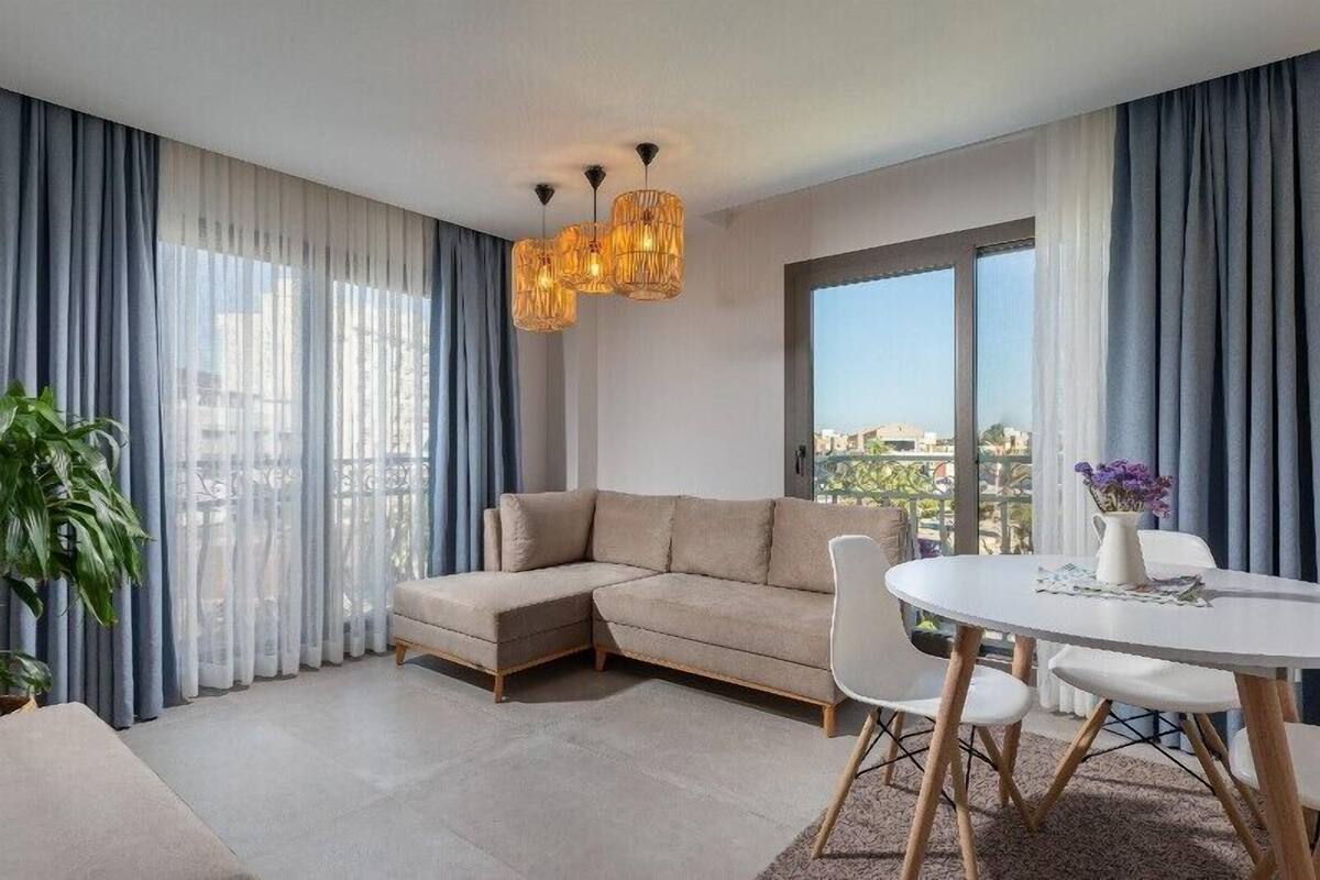 Een moderne woonkamer met een hoekbank, een ronde eettafel en een groot raam met gordijnen.