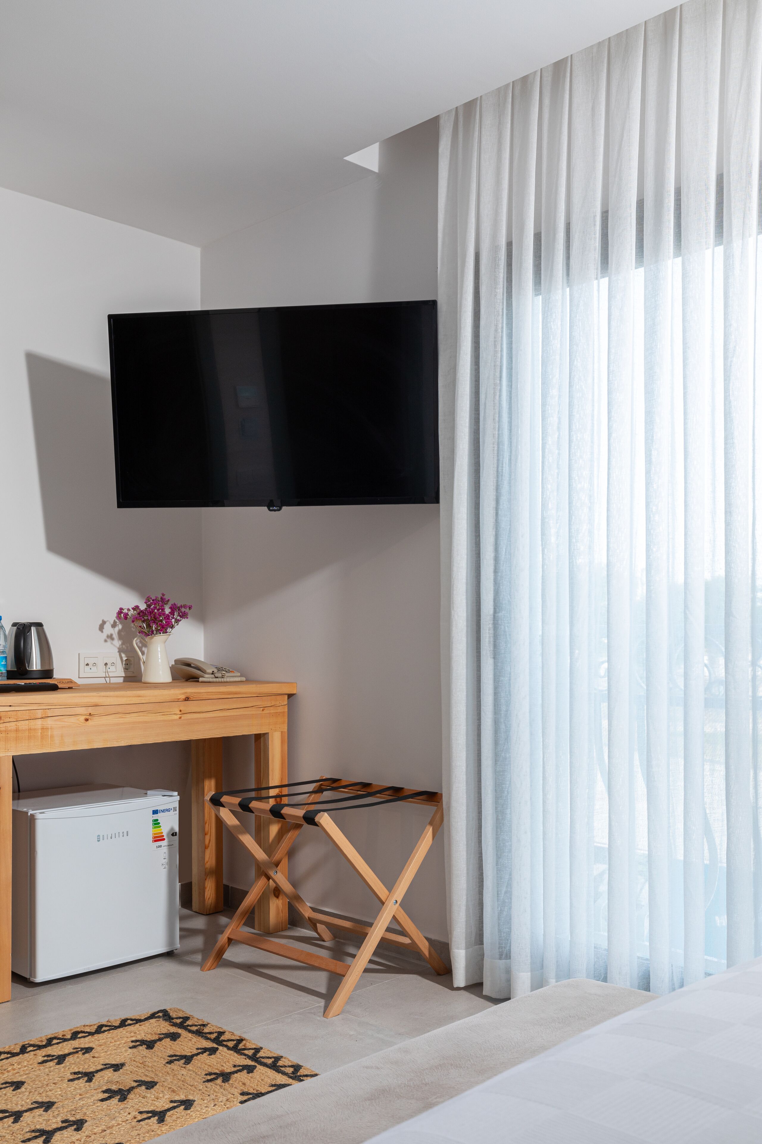 A hotel room with a flat-screen TV mounted on the wall, a wooden desk with a small refrigerator, a telephone, a vase with flowers, and a window with sheer curtains.
