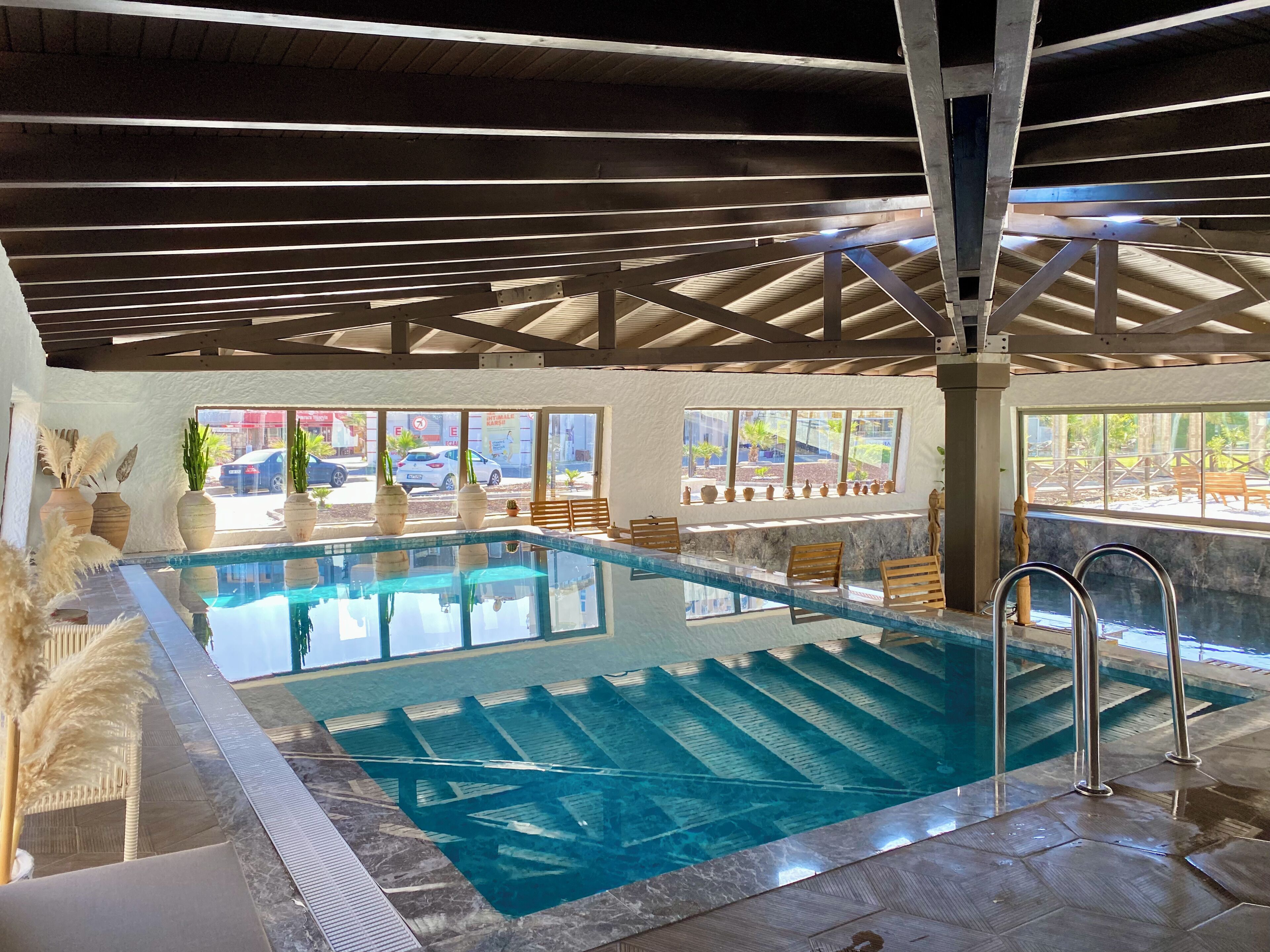 An indoor swimming pool with clear water, wooden deck chairs, and large windows offering views of the outside.
