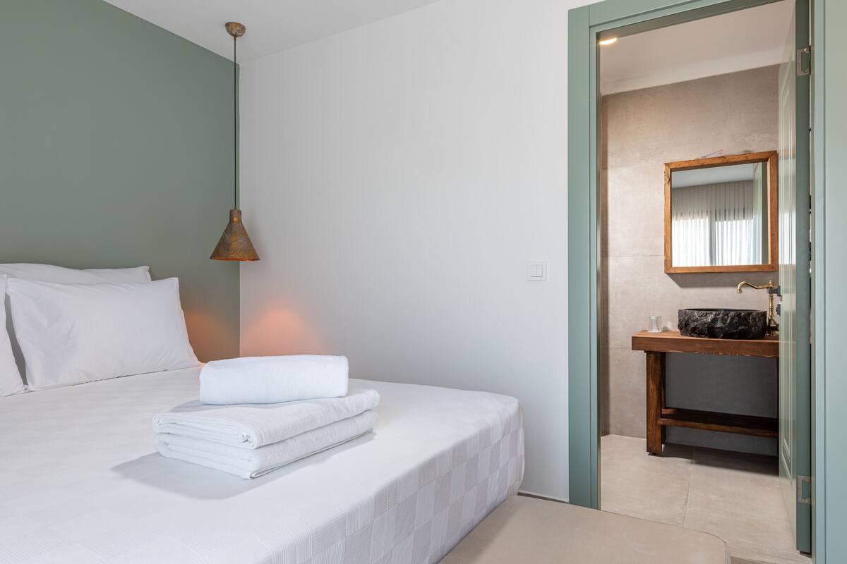Een hotelkamer met een opgemaakt bed, een hangende hanglamp en een badkamer met wastafel en spiegel.