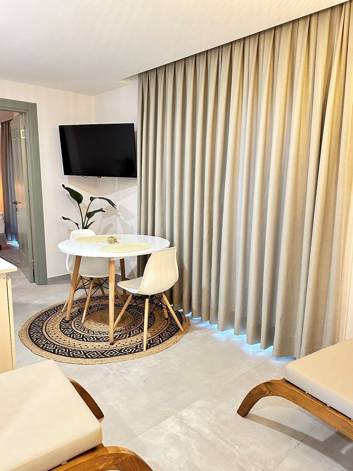 A modern hotel room with a round dining table, white chairs, a flat-screen TV mounted on the wall, and beige curtains.
