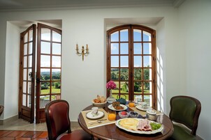 A dining area with a table set for breakfast, large windows with a view of the outdoors, and a chandelier on the wall.