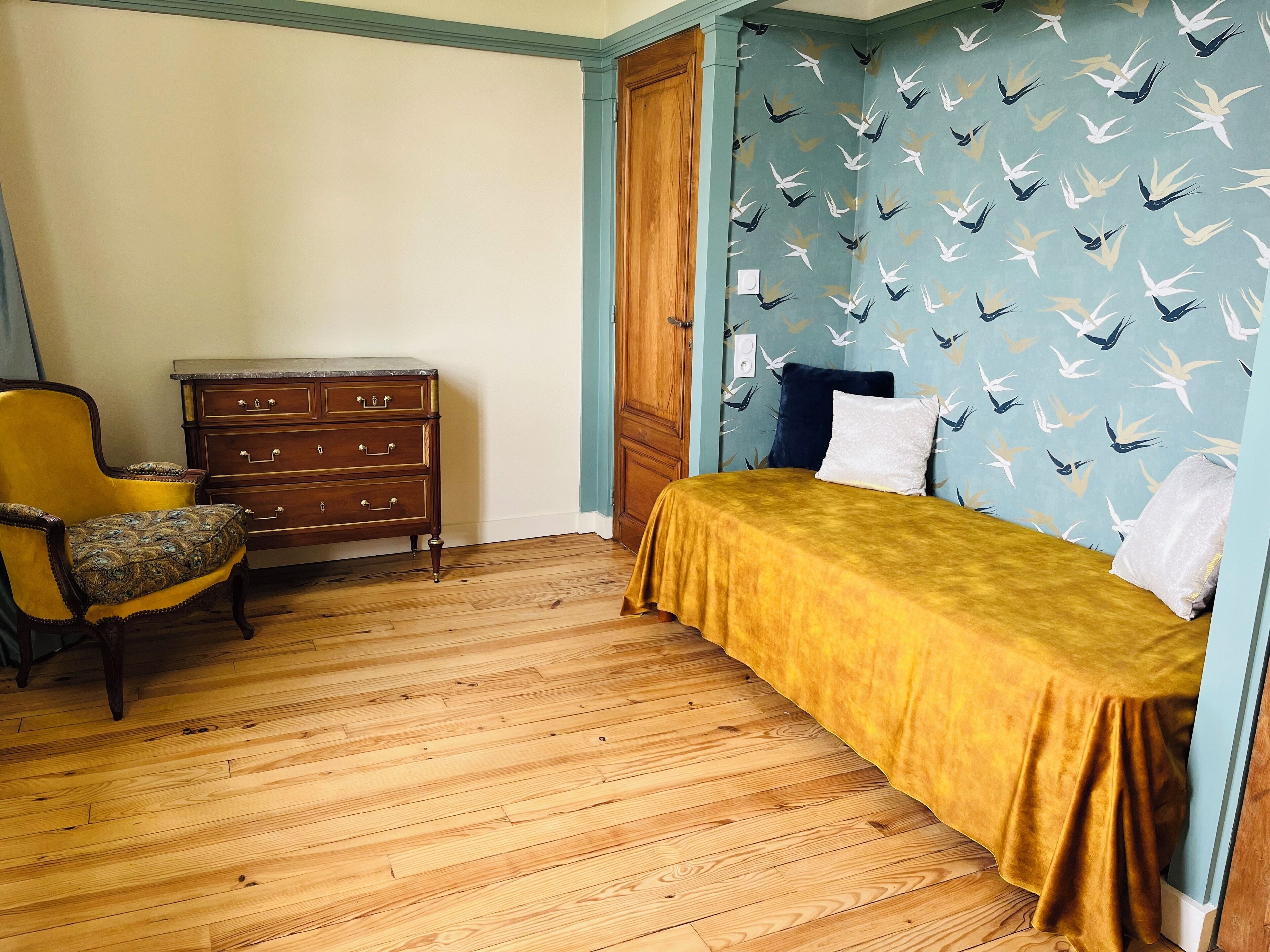 A room with a wooden floor, a bed with a yellow bedspread, a wooden dresser, a chair with a patterned cushion, and a wall with a bird pattern.
