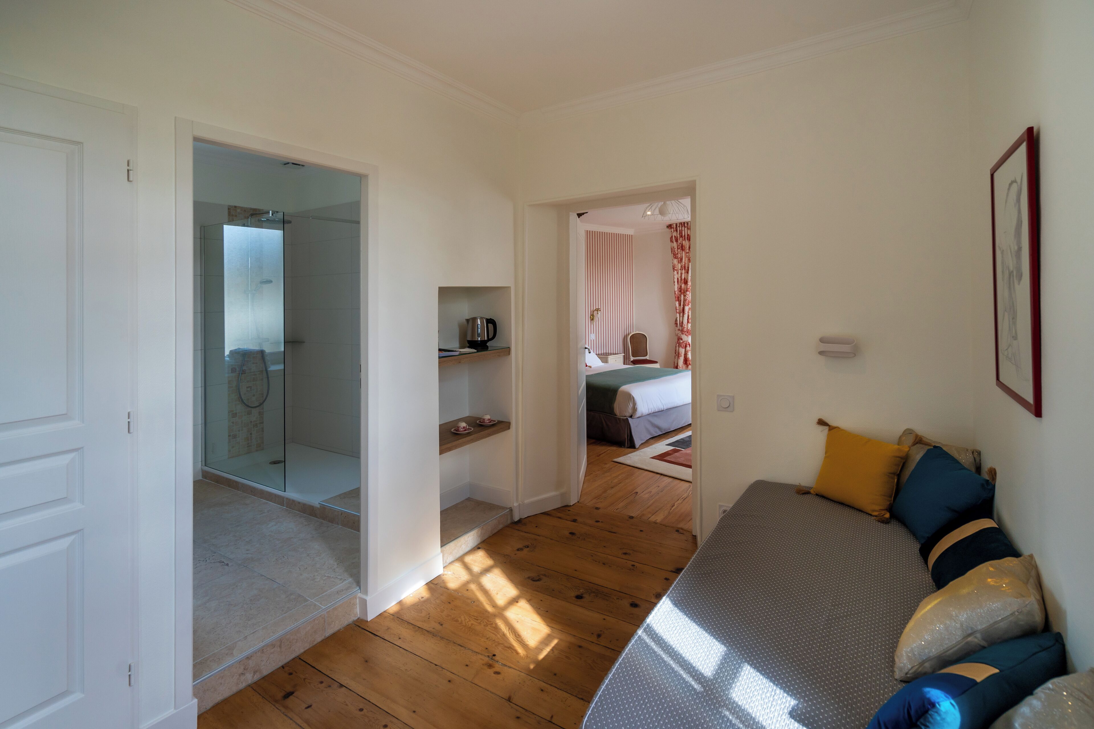 A bedroom with a bed, wooden flooring, a shelf with a kettle, and a bathroom with a shower.