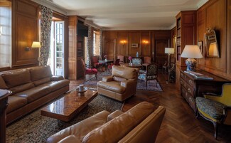 A spacious living room with leather sofas, a coffee table, and a fireplace. There are wooden panel walls and a large window with curtains.