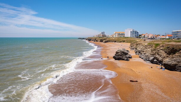 A beach with clear skies, rocky outcrops, and buildings in the background.