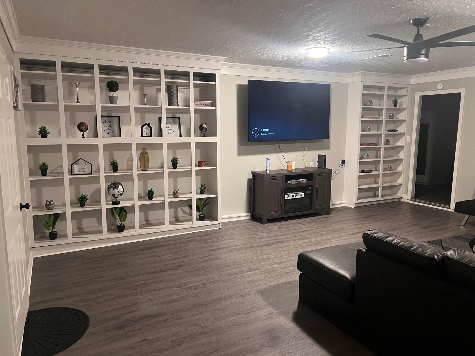 A living room with a flat-screen TV mounted on the wall, a dark wooden entertainment unit, and a black leather sofa.