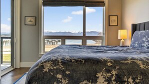Une chambre avec une grande fenêtre, un lit recouvert d’une couvre-lit à motifs, une table de chevet avec une lampe et des tableaux encadrés accrochés au mur.
