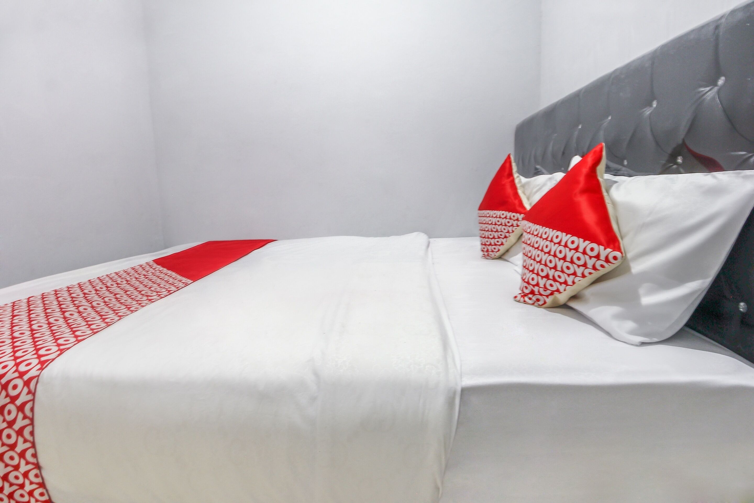 Una cama bien hecha con ropa blanca y roja, un cabecero tapizado en gris y una almohada roja y blanca con un estampado repetido.