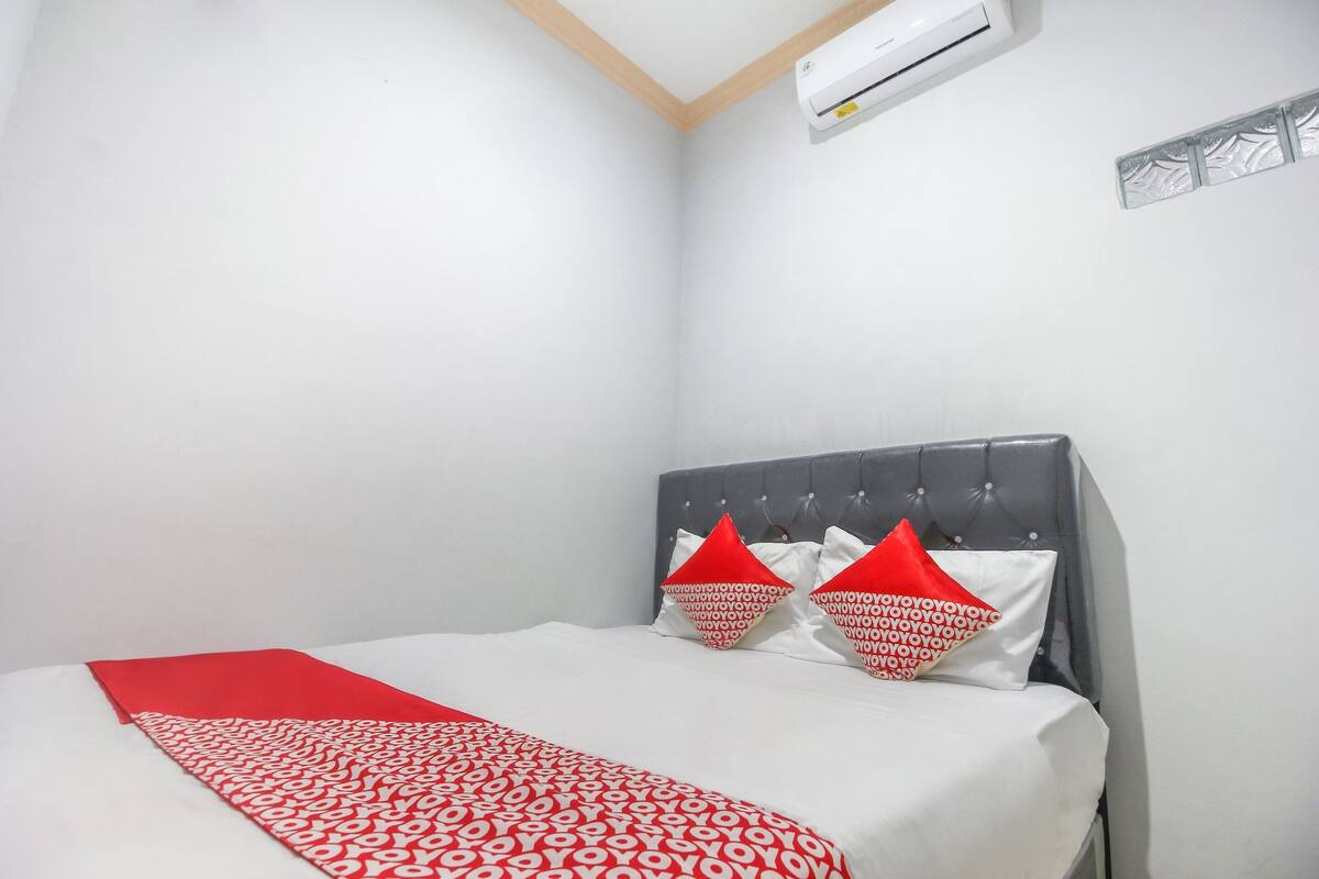 Cama bem arrumada com colcha estampada em vermelho e branco, cabeceira estofada cinza e duas almofadas vermelhas com texto branco.