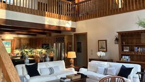 A spacious living area with a high ceiling, wooden beams, a large window, and a kitchen in the background. - Lakefront, 4Bdr+Loft, 3Ba, Boat Ramp, Golf, Ping Pong, Foosball, No Pets (Drasco)