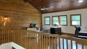 A spacious room with wooden walls and ceiling, a ceiling fan, a television, a sofa, a dresser, and three windows with a view of greenery. - Lakefront, 4Bdr+Loft, 3Ba, Boat Ramp, Golf, Ping Pong, Foosball, No Pets (Drasco)