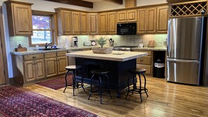 A modern kitchen with wooden cabinets, a central island with stools, stainless steel appliances, and a red patterned rug. - Lakefront, 4Bdr+Loft, 3Ba, Boat Ramp, Golf, Ping Pong, Foosball, No Pets (Drasco)