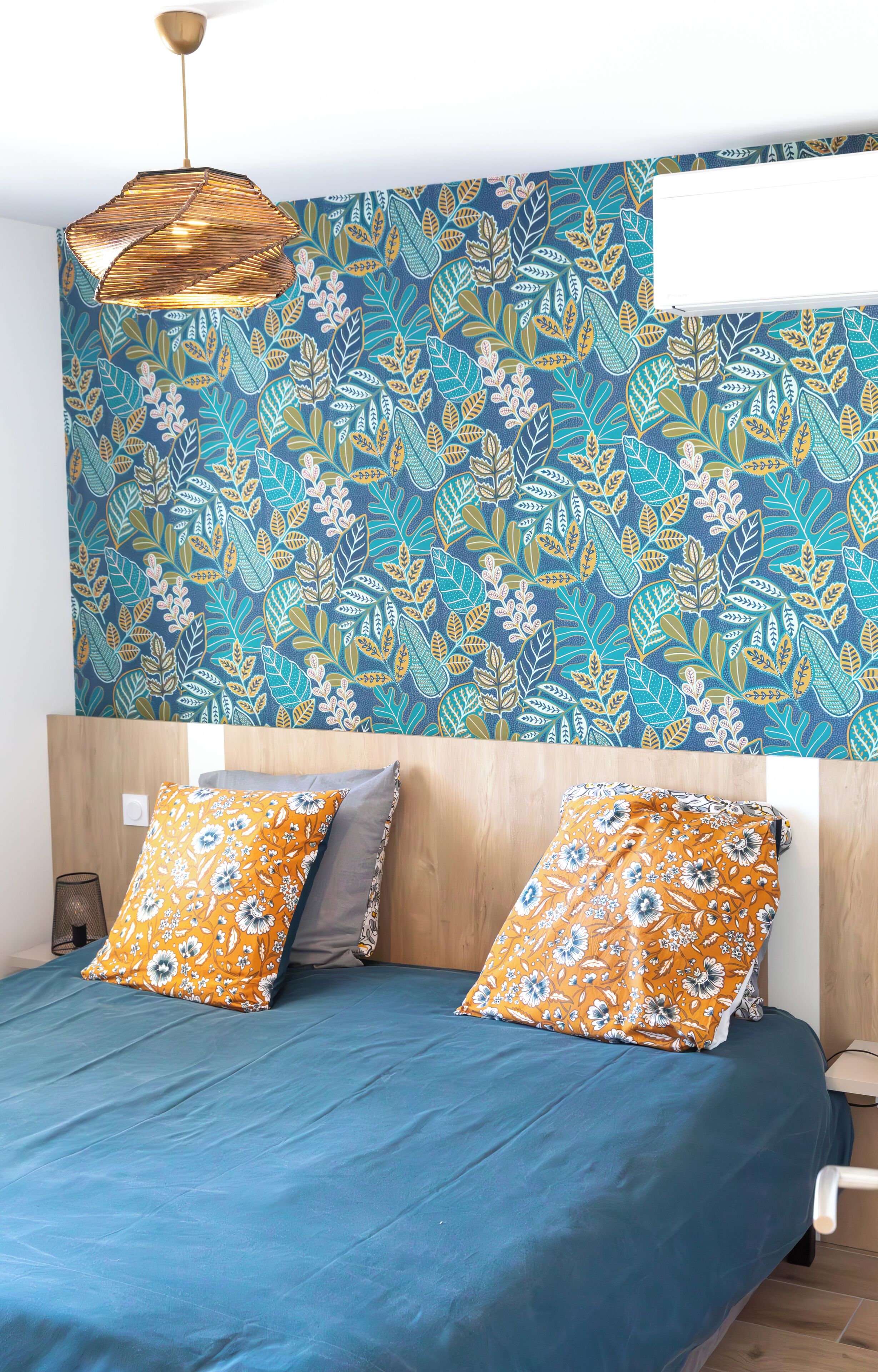 A bedroom with a blue bedspread, two orange floral pillows, a wooden headboard, and a wall with a leafy pattern.