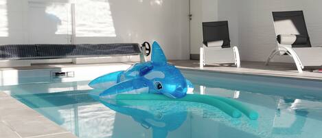 A swimming pool with a blue shark float, tiled walls, and two white lounge chairs.